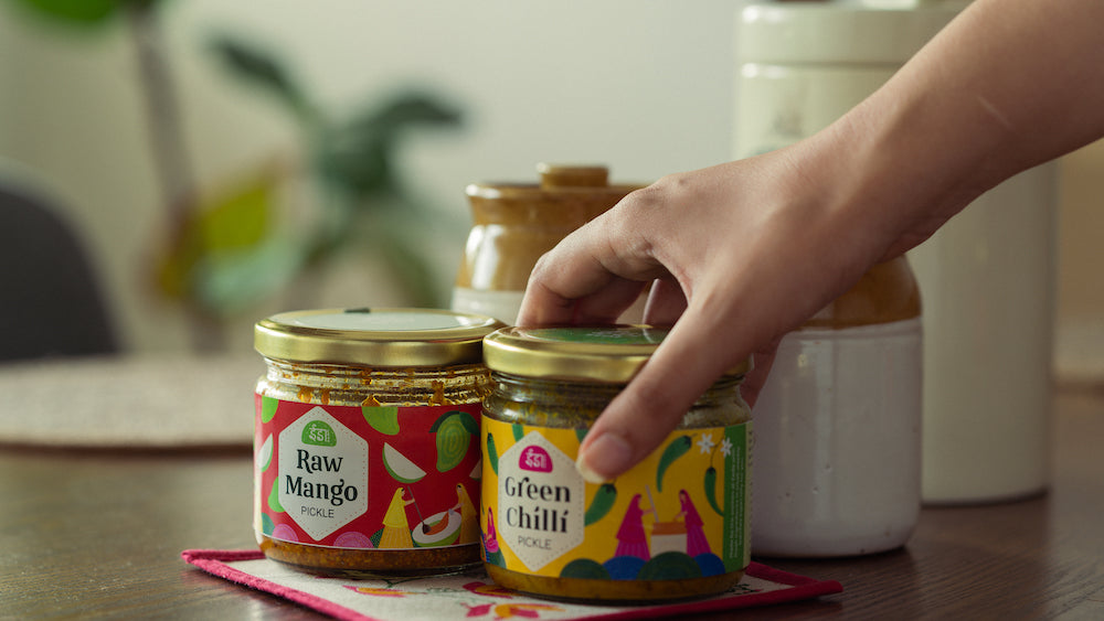 A hand reaching for jars of Eeda Raw Mango and Green Chilli pickle on a kitchen table